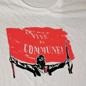 Może przedstawiać: Biała koszulka z czerwoną flagą i czarną sylwetką osoby trzymającej karabin. Na fladze widnieje napis "Vive la Commune!"