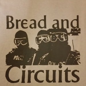 Puede incluir: Una camiseta gris con un estampado gráfico negro de tres figuras con pasamontañas y gafas de esquí. El texto "Bread and Circuits" está impreso debajo de las figuras.