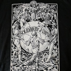 Pode incluir: Estampa gráfica em preto e branco em uma camiseta preta. O gráfico representa um globo com figuras de mãos dadas ao redor dele. O texto "Solidariedade do Trabalho" está escrito no globo. As figuras representam diferentes continentes: África, Ásia, América, Austrália e Europa.