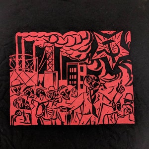 Peut inclure: Impression rouge et noire sur un t-shirt noir représentant une foule de personnes devant une usine avec de la fumée qui s'échappe de la cheminée. Une étoile rouge avec un marteau et une faucille se trouve dans le coin supérieur droit.