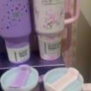 May include: A lavender tumbler with a dotted pattern and a white tumbler featuring pink text and graphics, both with matching light blue lids. Each tumbler has a handle and a sliding closure on the lid. The white tumbler displays the text "Stay Wild".