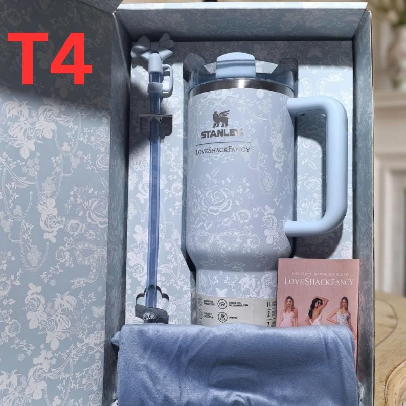 May include: A light blue Stanley tumbler with a white floral pattern and the LoveShackFancy logo, featuring a matching straw with a bow topper, presented in a decorative box with a small informational card and a folded blue fabric accessory.