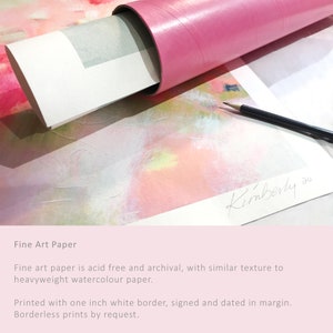 May include: A close-up of a fine art print rolled up in a pink tube. The print is a watercolor painting with pink, yellow, and white colors. The print is signed "Kimberly" in the bottom right corner. The text "Fine Art Paper" is visible at the bottom of the image. The text describes the paper as acid-free and archival, with a similar texture to heavyweight watercolor paper. The print is printed with a one-inch white border, signed and dated in the margin. Borderless prints are available by request.