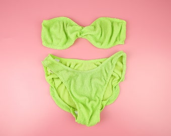 Könnte beinhalten: Ein limegrüner Bikini-Oberteil und -Unterteil mit gerippter Textur. Das Oberteil ist ein Bandeau-Stil mit einem Knoten vorne. Das Unterteil ist ein hoch taillierter Stil mit einem Rüschenrand.