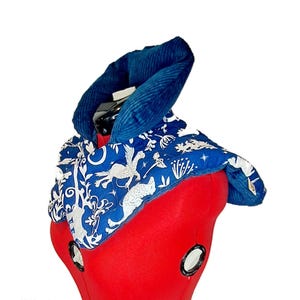 May include: A neck wrap featuring a deep blue corduroy collar and a blue fabric body with a white repeating pattern of mythical creatures, stars, and floral elements. The quilted accessory is draped over a red mannequin form for display.