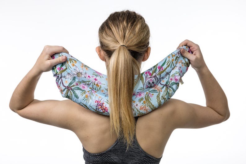 May include: A light blue neck wrap featuring a colorful botanical print with pink flowers, green leaves, and small grey koala illustrations. The fabric accessory is shaped to drape comfortably around the neck and shoulders for support.