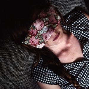 May include: A rectangular eye mask featuring a dark brown background with a dense pattern of pink and white roses. The mask is positioned over the eyes, paired with a black and white gingham patterned top with small floral accents on the fabric.