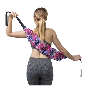 May include: A long, cylindrical fabric yoga mat carrier featuring a vibrant floral pattern in shades of pink, purple, blue, and cream. The carrier includes a long, adjustable black strap that extends across the back for carrying over the shoulder.
