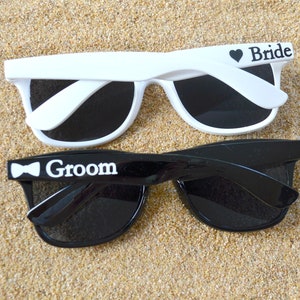 May include: Two pairs of sunglasses with black lenses rest on sand, one white and one black. The white sunglasses have "Bride" printed on the arm with a heart, and the black sunglasses have "Groom" printed with a bow tie. Seashells are scattered around.