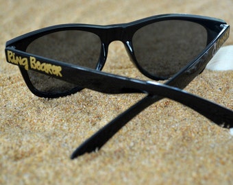 May include: Black sunglasses with dark lenses featuring the text Ring Bearer printed in gold on the left temple and the name Connor with a small ring icon printed in gold on the right temple, resting on a textured sandy surface.