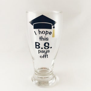 May include: A clear glass beer mug with a black graduation cap and tassel design. The mug has the text "I hope this B.S. pays off!" printed on it.