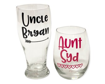 May include: Two clear glass beverage glasses with personalized text. The larger glass has "Uncle Bryan" with a black arrow pointing to the right. The smaller glass has "Aunt Syd" in pink text with a row of pink hearts at the bottom.