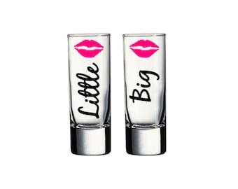 May include: Two clear glass shot glasses with pink lips printed on the front. One glass says "Big" and the other says "Little".