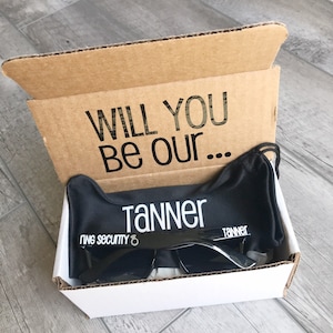 May include: A cardboard box with the text "WILL YOU BE OUR..." printed on the inside flap. Inside the box is a black sunglasses case with the name "TANNER" and the words "RING SECURITY" printed on it. Black sunglasses are inside the case.