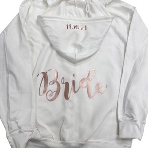 May include: White hooded sweatshirt with the word "Bride" in rose gold script across the back. The date "11.16.21" is printed in rose gold on the hood. The sweatshirt has a drawstring and ribbed cuffs.