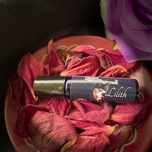 May include: A blue glass roll-on bottle with a black cap, labeled "The Garden of earthly Delights" and "Lilith". The bottle is surrounded by red rose petals in a red ceramic dish.