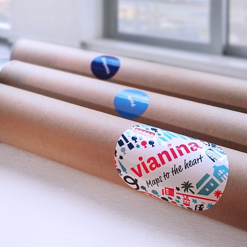 Op de afbeelding: Drie bruine kartonnen buizen met kleurrijke ronde stickers. De sticker op de onderste buis heeft een witte achtergrond met een kleurrijk ontwerp en de tekst "vianina Maps to the heart".