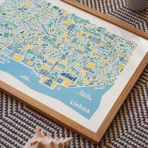 May include: A framed art print of Lisbon, Portugal, featuring a detailed, colorful map illustration. The map uses blue, yellow, and green hues to depict the city's layout, with the word "Lisboa" printed in white. The print is set in a natural wood frame.