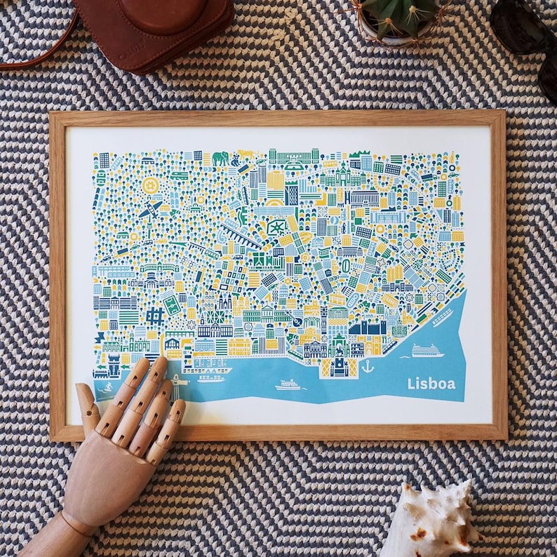 May include: A framed art print of a colorful map of Lisbon, Portugal. The map features a blue, yellow, and green color scheme with detailed illustrations of buildings and landmarks. The word "Lisboa" is printed in white on the bottom right.