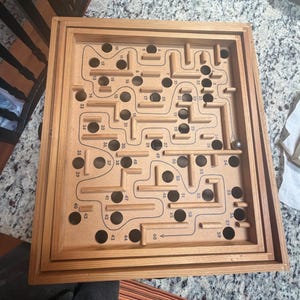 May include: A rectangular wooden labyrinth game featuring a light brown board with raised wooden walls, numerous circular holes, and a metal ball. The board is printed with a winding path and numbered positions from 1 to 60, guiding the ball through the maze.