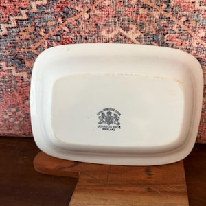May include: A rectangular, off-white ceramic dish with rounded corners. The underside features a black and white emblem with the text "Royal Ironstone China" and "Johnson Bros England". The dish rests on a wooden cutting board.