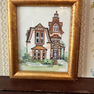 May include: A framed watercolor painting of a Victorian house in a gold frame. The house is white with brown trim and a brown roof. The text "Georgetown Colorado" is visible at the bottom of the painting.