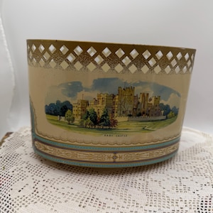 May include: A vintage beige container with a diamond-patterned top edge and a painted image of Raby Castle. The container has a light blue and gold trim. The image is set on a white lace doily.