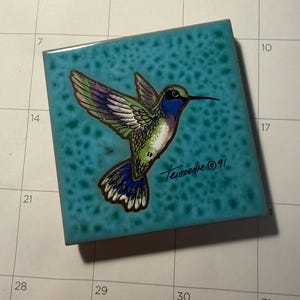 May include: A square ceramic tile with a glossy turquoise background featuring a speckled pattern. A hummingbird with green, blue, and purple feathers is depicted in flight. The signature Teissedre and the year 91 are written in black ink.
