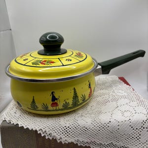 May include: A yellow saucepan with a lid and a dark green handle. The saucepan is decorated with a painted scene of figures and trees. The lid has a dark green knob and floral designs. The saucepan is sitting on a white lace doily.