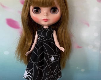 Puede incluir: Una muñeca con cabello castaño largo y flequillo, vestida con un vestido negro con un diseño de telaraña y araña blanco. La muñeca tiene mejillas rosadas y usa botas negras con detalles rojos. El fondo presenta un diseño floral.