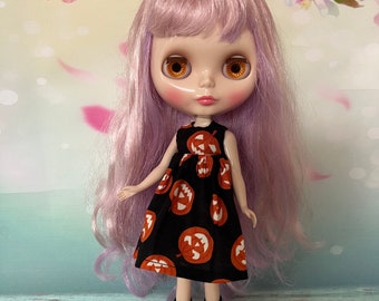 Puede incluir: Una muñeca con cabello lavanda y ojos grandes y anaranjados. La muñeca viste un vestido negro con calabazas naranjas y botas negras con cordones rojos. El fondo presenta un diseño floral con pétalos rosas y blancos.