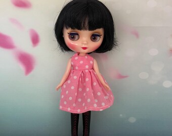 May include: A doll with black hair and a pink dress with white polka dots. The doll is wearing black boots with red soles. The background is a soft gradient of blue and pink with floral accents.