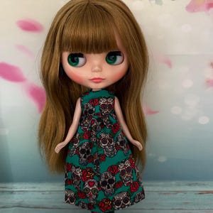 May include: A doll with long, light brown hair and green eyes wearing a teal dress. The dress is patterned with sugar skulls and red roses. The doll is wearing black boots and has rosy cheeks.