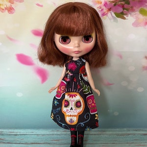 May include: A doll with brown hair and pink eyes wearing a black dress with a colorful sugar skull design. The dress features floral and decorative elements. The doll is also wearing black boots.