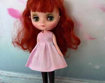 May include: A doll with red hair, green eyes, and freckles, wearing a light pink dress and black boots with red soles. The doll has a large head and is posed against a light blue and pink background.