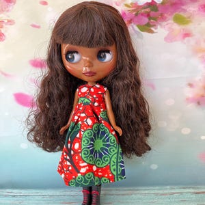 May include: A doll with brown hair and blue eyes wearing a red, green, and white patterned dress and black boots. The doll has a fair complexion and is posed against a floral background.