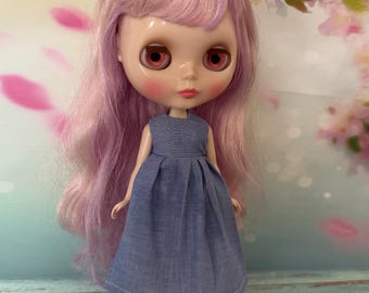 May include: A doll with long, wavy pastel pink hair, large eyes with reddish irises, and rosy cheeks. The doll wears a sleeveless blue chambray dress with a pleated skirt and black boots with red laces. The doll has a pale complexion.