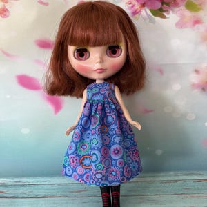 May include: A doll with auburn hair and large, pink-rimmed eyes. The doll is dressed in a blue dress with colourful floral patterns and black boots with red detailing. The background features a floral design with pink and white blossoms.