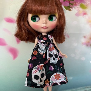 May include: A doll with auburn hair and green eyes wearing a black dress. The dress is patterned with white sugar skulls, flowers, and spiderwebs. The doll has rosy cheeks and a neutral expression.