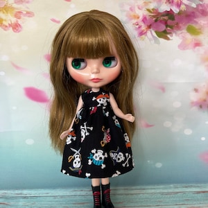 May include: A doll with long brown hair and green eyes wearing a black dress with a skull and crossbones pattern. The doll is also wearing black boots with red laces. The background has a floral design.