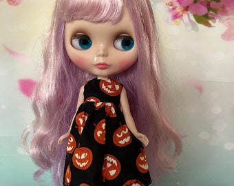 Puede incluir: Una muñeca con pelo largo y ondulado color lavanda y grandes ojos azules. La muñeca lleva un vestido negro con un estampado de caras de calabaza naranja. Las mejillas de la muñeca son rosadas y el fondo es un diseño floral suave.
