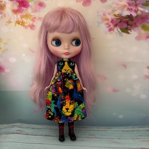 May include: A doll with long, wavy pastel pink hair and blue eyes. The doll wears a sleeveless dress featuring a vibrant, multicolored cat face print against a black background, paired with black boots that have red laces and soles.