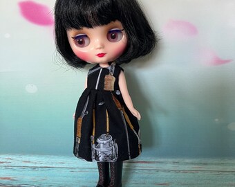 May include: A doll with black hair and large eyes wearing a black dress with a pattern of tools and a lantern. The doll is also wearing black boots with red soles. The background is a light green and pink.