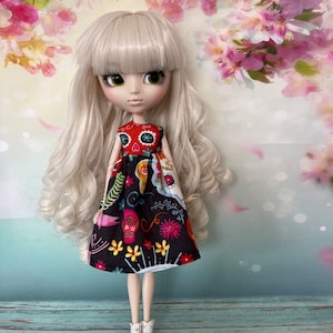 May include: A doll with long, wavy, light blonde hair and green eyes, wearing a black dress with a colorful Day of the Dead skull and floral pattern. The doll is wearing gray and white socks. The background features a floral design.