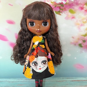 May include: A doll with dark skin, brown hair, and large eyes. The doll wears a black dress with a colorful Day of the Dead design, featuring a skull face. The dress has yellow and orange floral accents. The doll is wearing black boots.