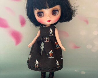 May include: A doll with black hair and a black dress featuring a pattern of skiers and trees. The doll has large eyes and is wearing black boots with red soles. The dress has a round neckline and a flared skirt.