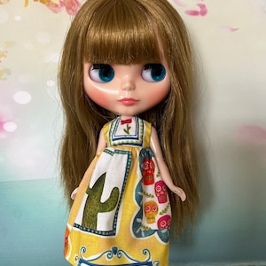 May include: A doll with long, light brown hair and blue eyes wearing a yellow dress with a cactus and skull print. The dress has a white and blue border. The doll has a fair complexion and is standing in front of a floral background.