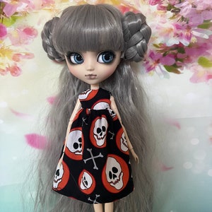 May include: A doll with gray hair styled in two buns and long, flowing strands. The doll wears a black dress with a pattern of white skulls inside orange circles and white crossbones. The doll has blue eyes and pale skin.