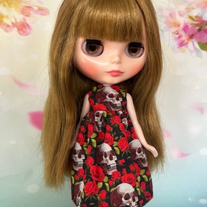 May include: A doll with long, light brown hair and a black dress patterned with white skulls and red roses. The doll has large eyes and is wearing a dress with a round neckline.