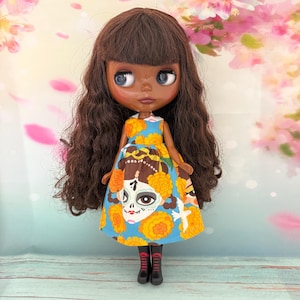 May include: A Blythe doll with dark skin, brown curly hair, and blue eyes. The doll wears a blue dress with a yellow floral pattern and a sugar skull design. The doll is wearing black boots.